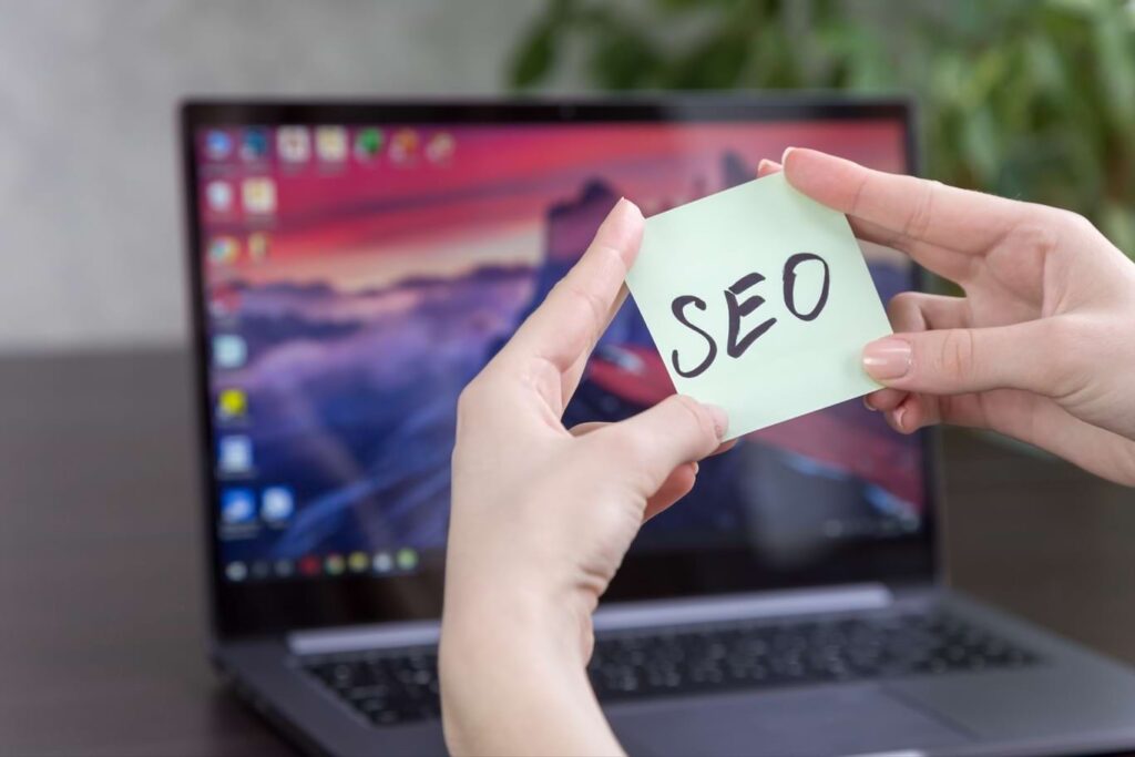 Manos de mujer sujetando un post-it que dice ‘SEO’ frente a una computadora portátil MacBook